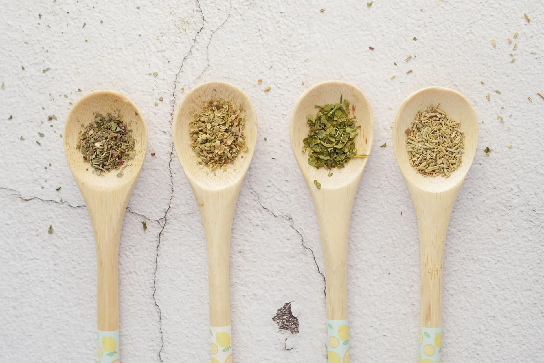 A photorealistic still life image of various preserved herbs: dried oregano hanging from a rustic string, frozen basil cubes in an ice tray with olive oil, and fresh rosemary sprigs stored in a glass jar with water. Cinematic lighting, sharp focus on the textures, depth of field, 8K, professional photography, shot on a high-end DSLR. Four wooden spoons with assorted dried herbs on a light background. — Foto: Towfiqu barbhuiya / Pexels